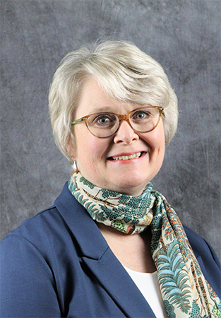 Professional portrait of a woman with short gray hair, glasses, and a blue blazer, wearing a patterned scarf, posed in front of a gray background.
