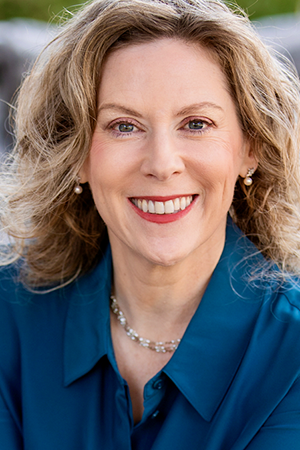 Professional portrait of a woman with wavy light brown hair, wearing a blue blouse and pearl earrings, smiling at the camera in an outdoor setting.