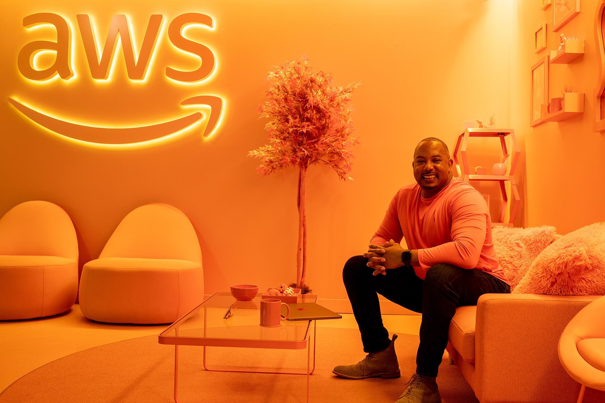 A man seated on a sofa in an orange-themed AWS office space, featuring modern furniture, decorative plants, and a prominent illuminated AWS logo on the wall.
