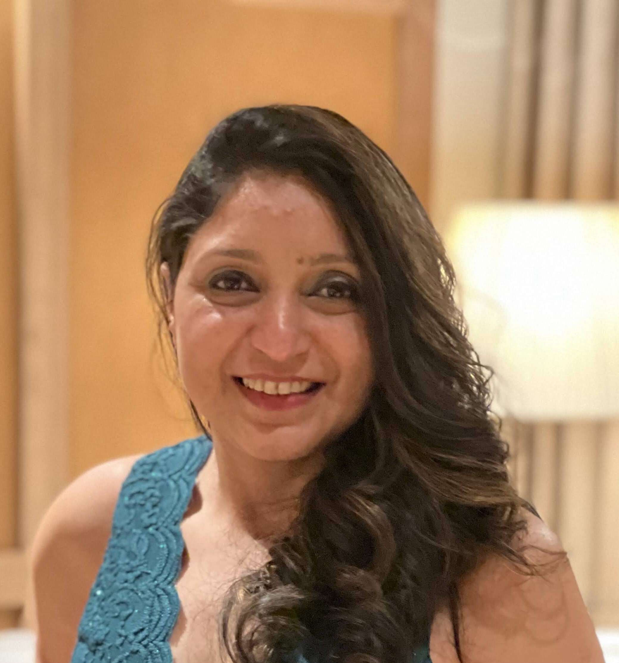 A portrait of a woman with long hair, smiling and wearing a teal dress, photographed indoors with a warm, softly blurred background.