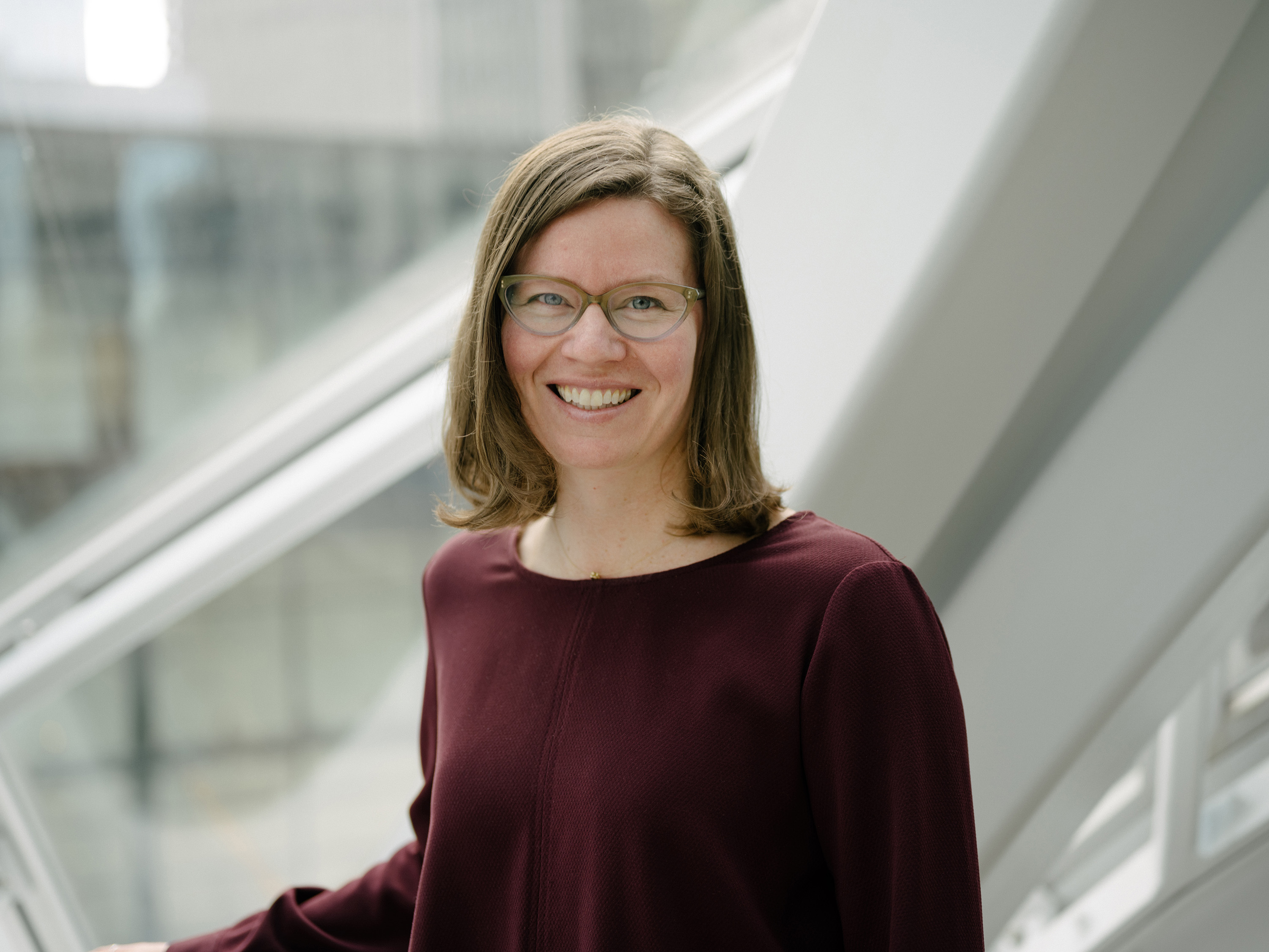 A professional portrait of a woman with glasses and shoulder-length hair, wearing a maroon top, smiling and standing in front of a modern glass and steel architectural background.