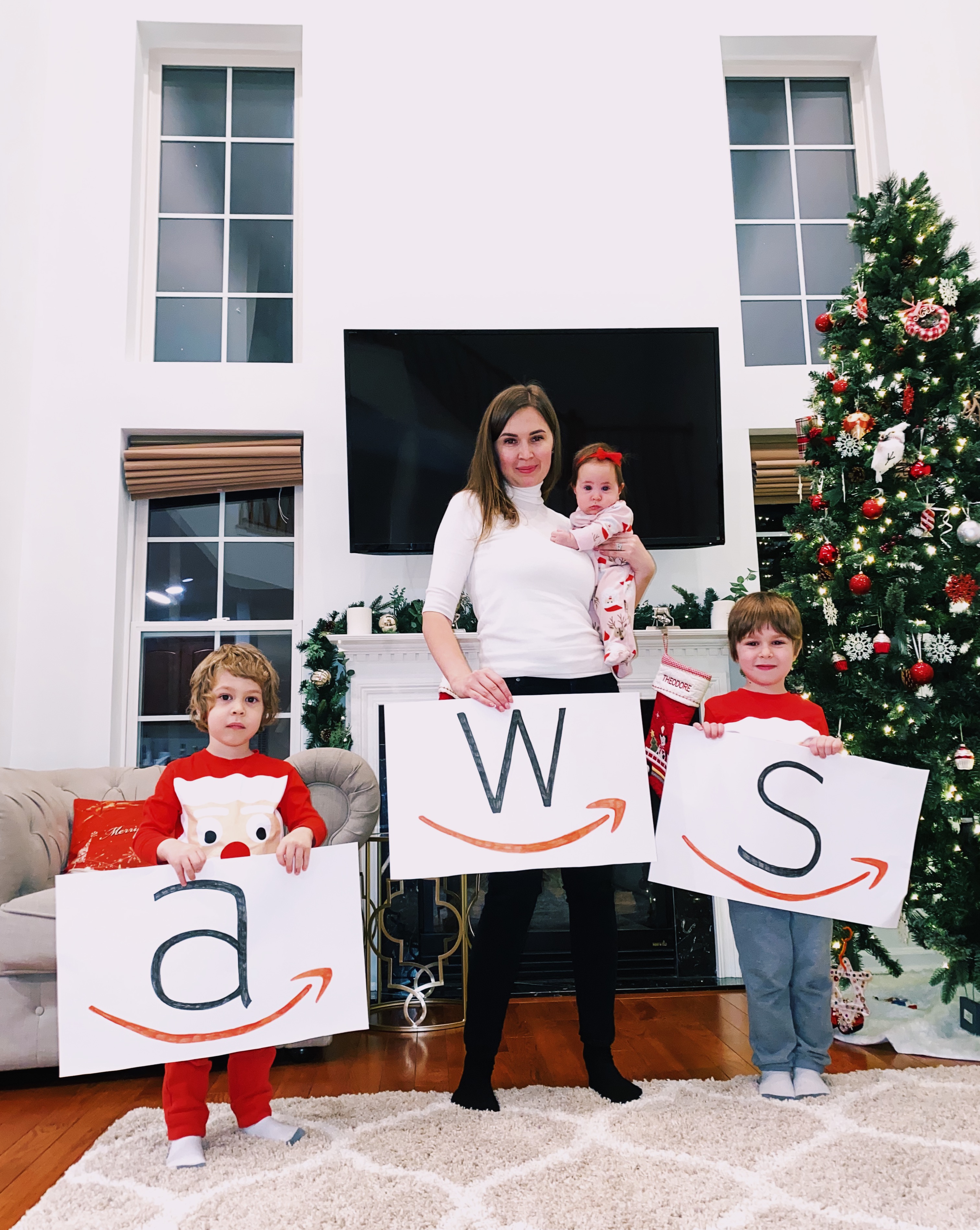A family stands together in a living room decorated for Christmas, holding large signs with the letters 'a', 'w', and 's'. A decorated Christmas tree is visible to the right, and festive decorations are present throughout the room.