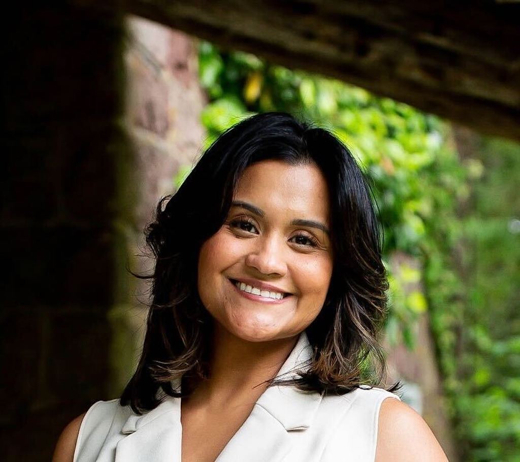 A woman with dark hair and a light sleeveless top smiling in an outdoor setting with greenery in the background.