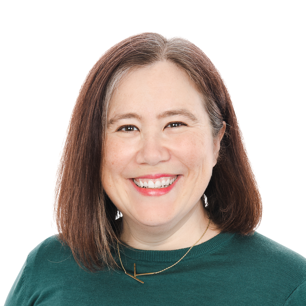 Headshot of a smiling woman with shoulder-length brown hair, wearing a green top and a necklace, on a plain background.