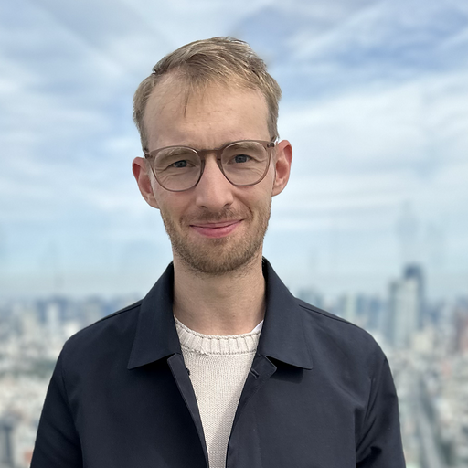 A man wearing glasses and a dark jacket stands in front of a blurred cityscape background.