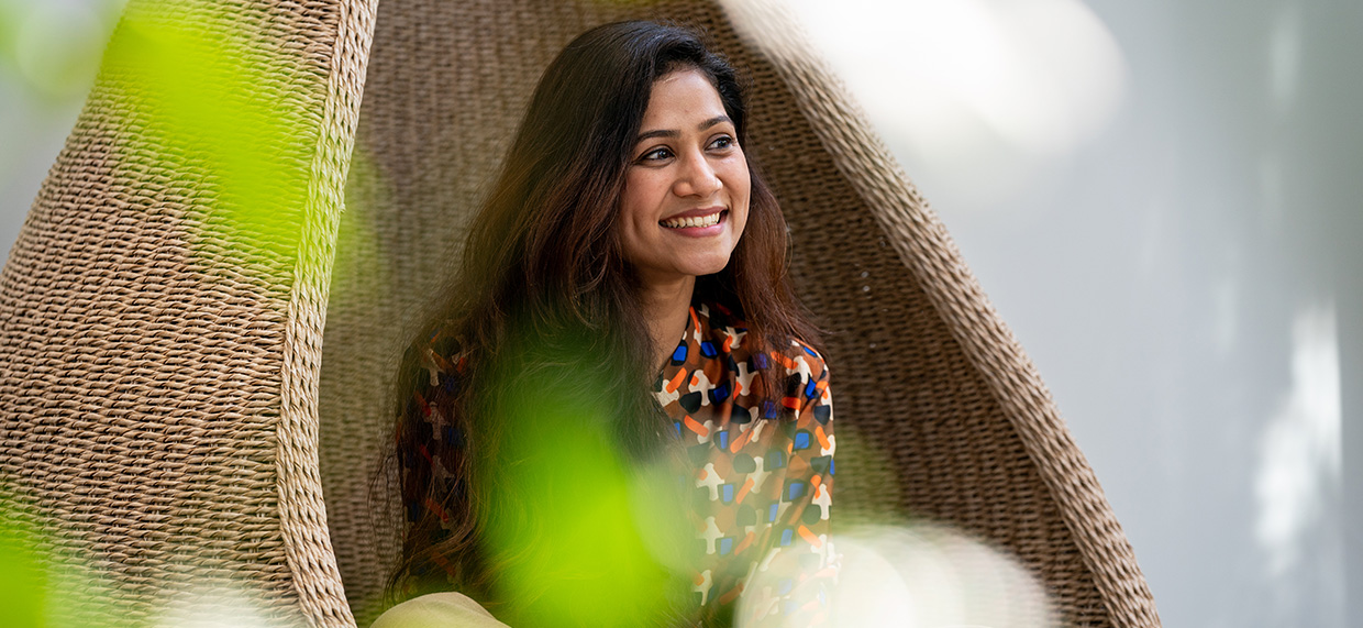 A smiling woman sits in a rounded wicker chair indoors with greenery in the foreground.
