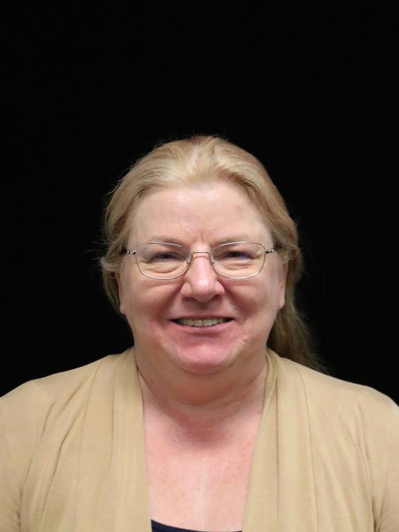 Portrait of a smiling woman with glasses and blonde hair, wearing a tan top, against a black background.