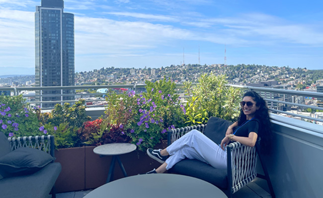 A woman wearing sunglasses and casual clothes is relaxing on a modern rooftop garden with colorful flowers and greenery, with a cityscape and tall building in the background under a blue sky.