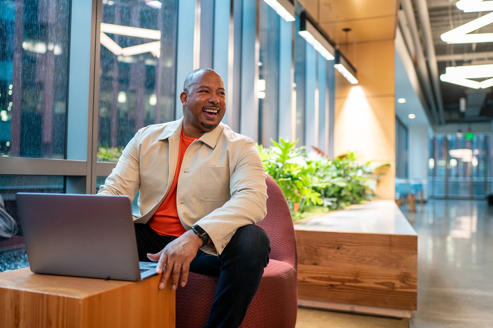 A man smiling while using a laptop, seated in a modern office lounge area with large windows and indoor plants.