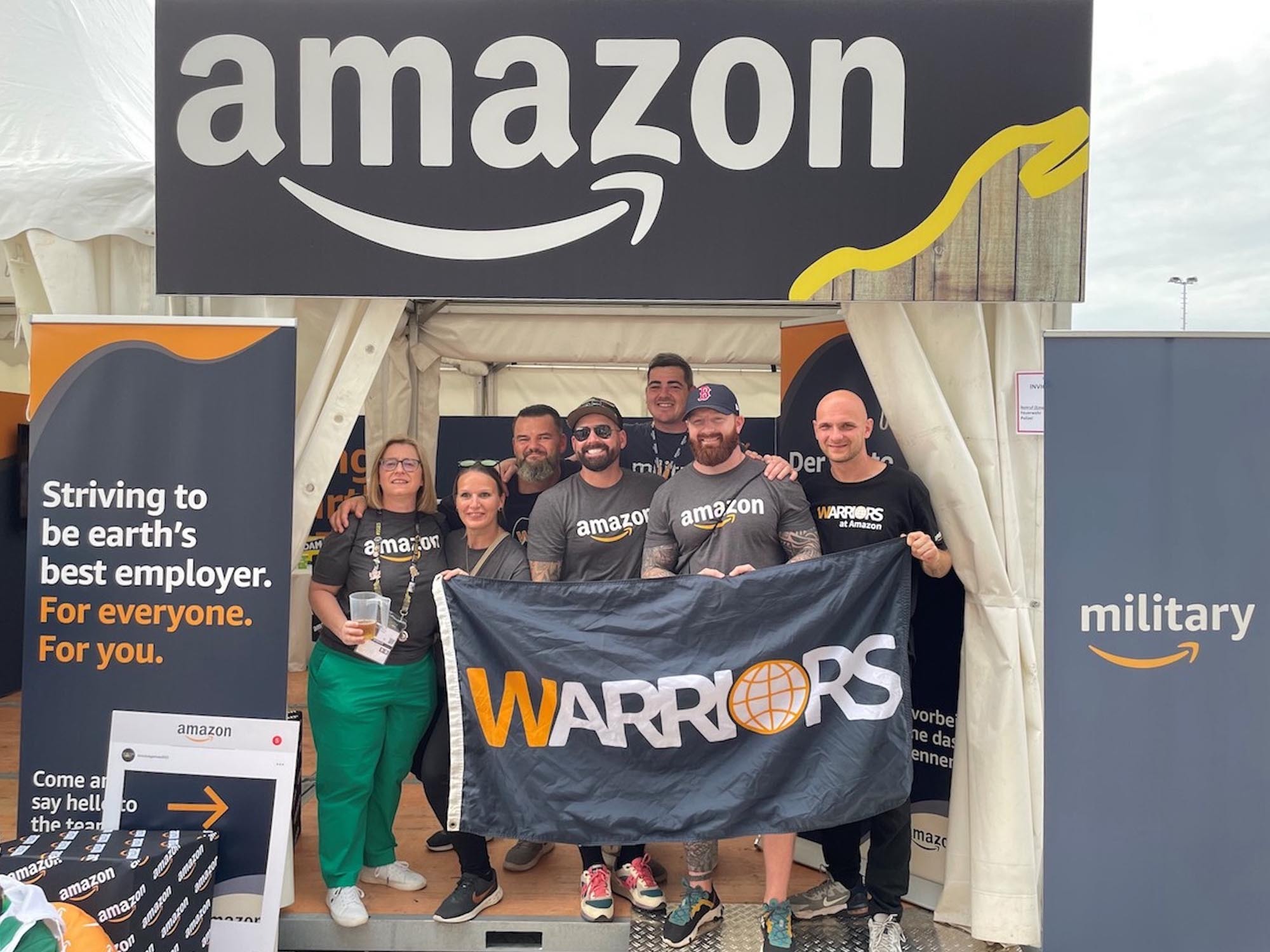 Group of Amazon employees standing together at a recruitment event tent, holding a 'Warriors' banner, with Amazon branding and military recruitment signage, promoting Amazon as an employer for everyone.