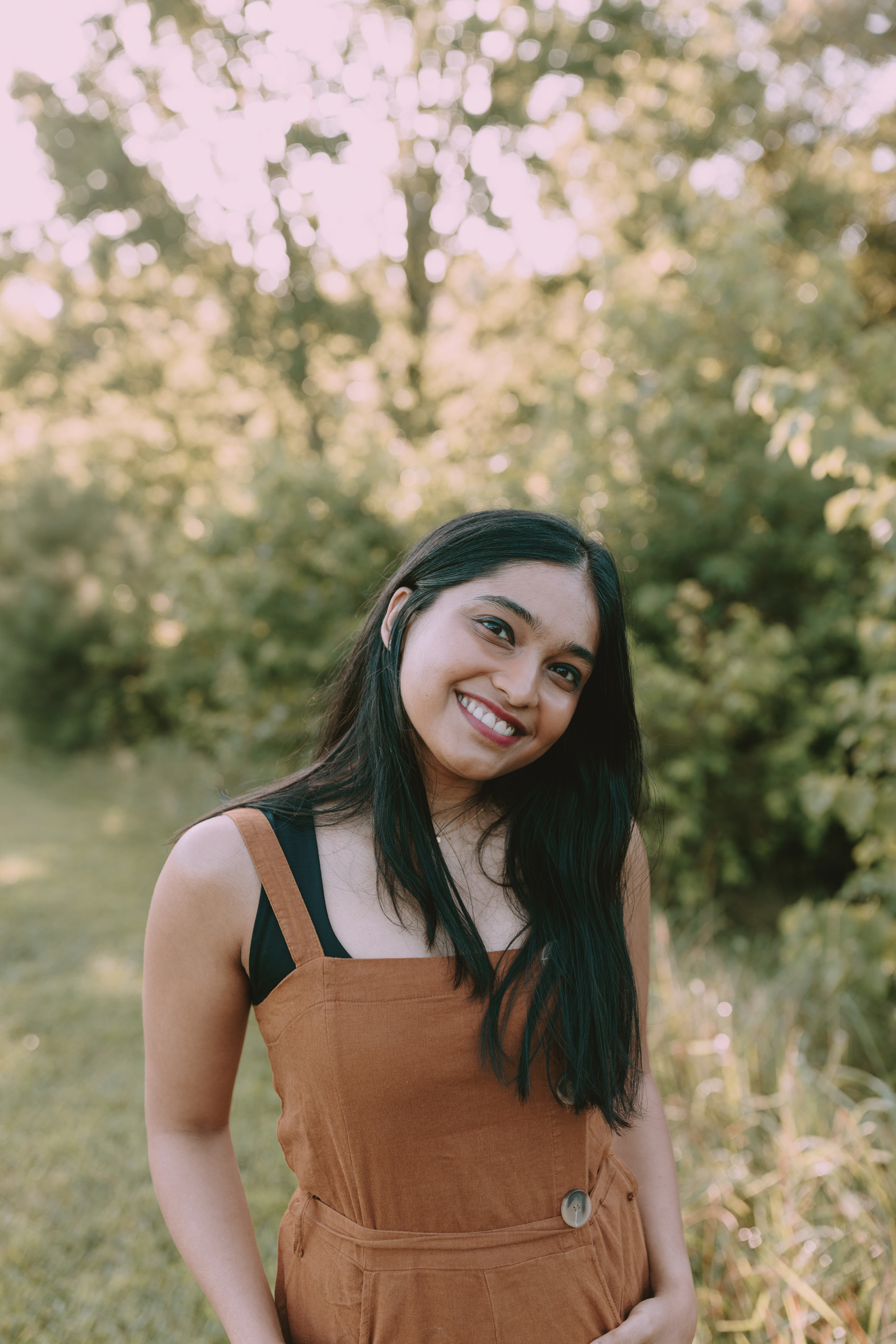 A woman smiling outdoors wearing a brown dress, standing in a lush green setting during summer.