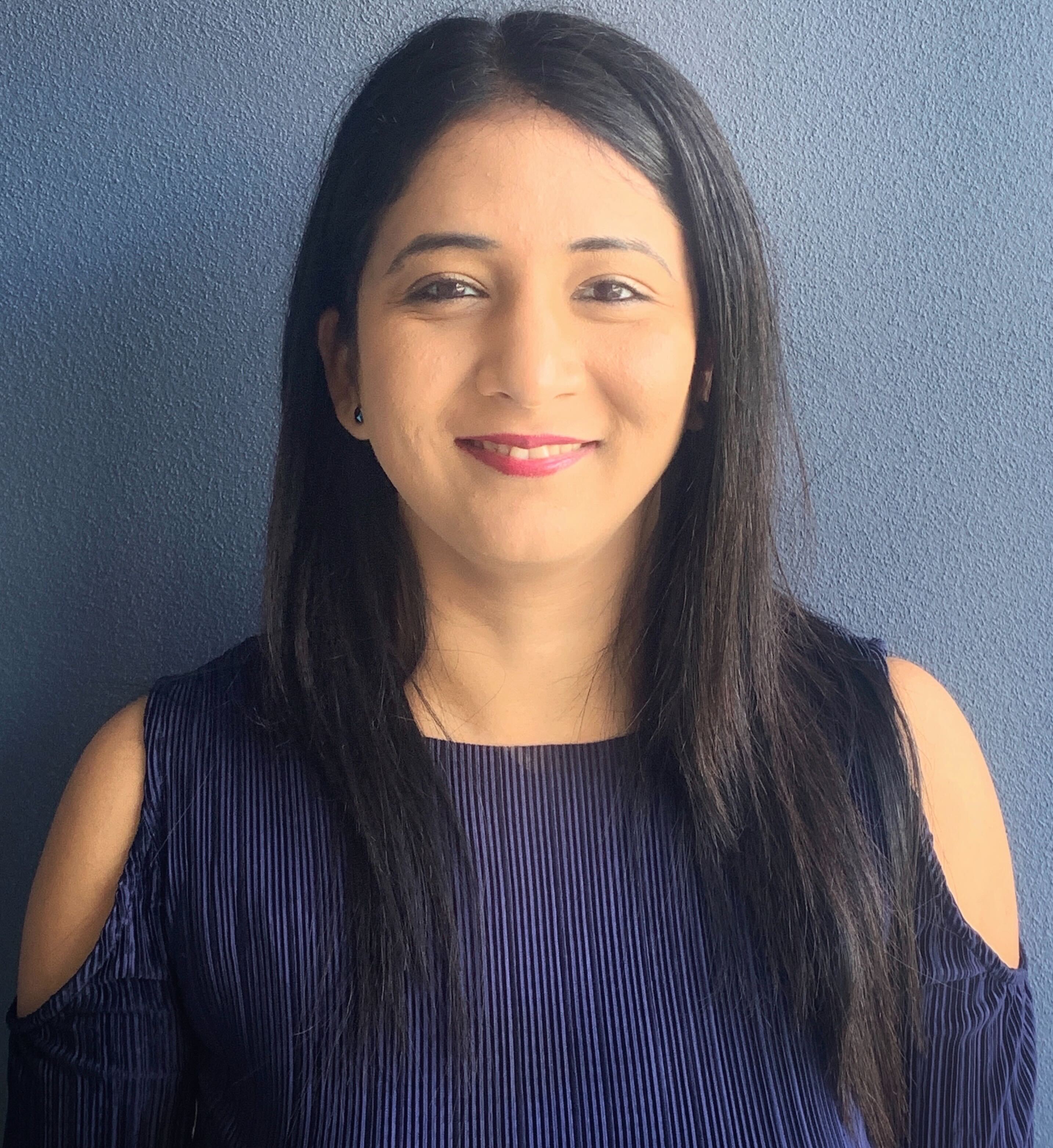 Portrait of a woman with long dark hair wearing a blue top, smiling, in front of a neutral background.
