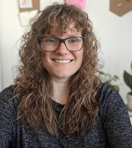A smiling woman with curly hair and glasses, wearing a dark top, is photographed indoors with a soft background featuring some plants and wall decor.