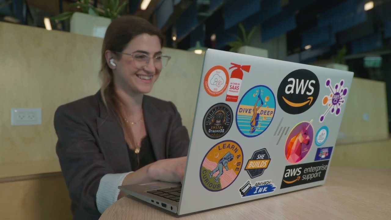 A woman smiling and using a laptop decorated with various AWS and technology-related stickers, seated in a modern workspace.