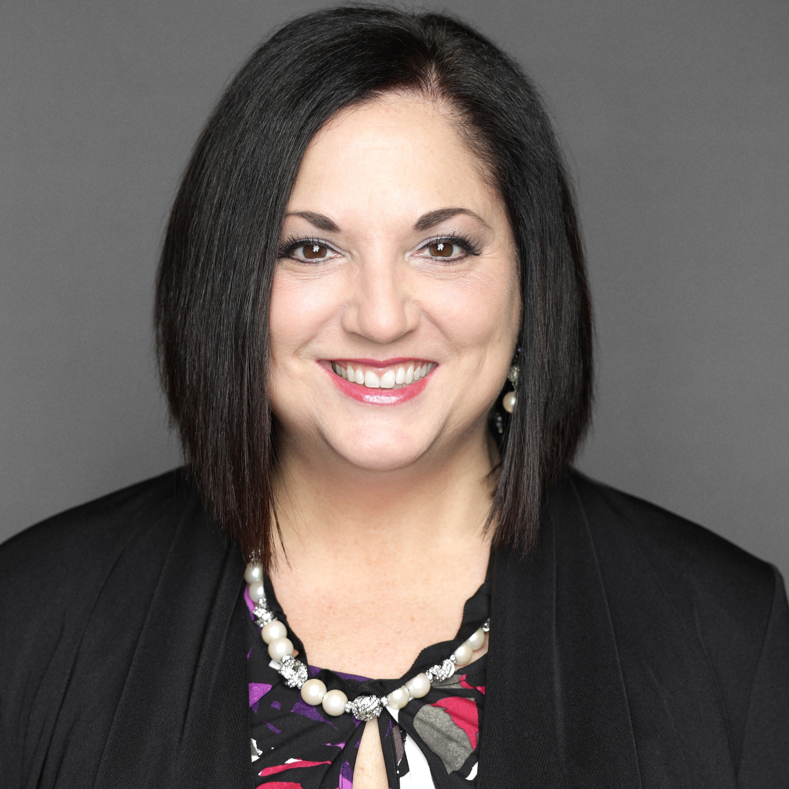 Professional headshot of a woman with dark hair, smiling, wearing a necklace and patterned blouse with a dark jacket, against a plain gray background.