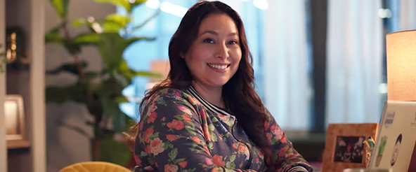 A woman wearing a floral jacket is smiling while sitting at a desk in an office setting. There is a plant, a desk lamp, and personal items visible in the background.