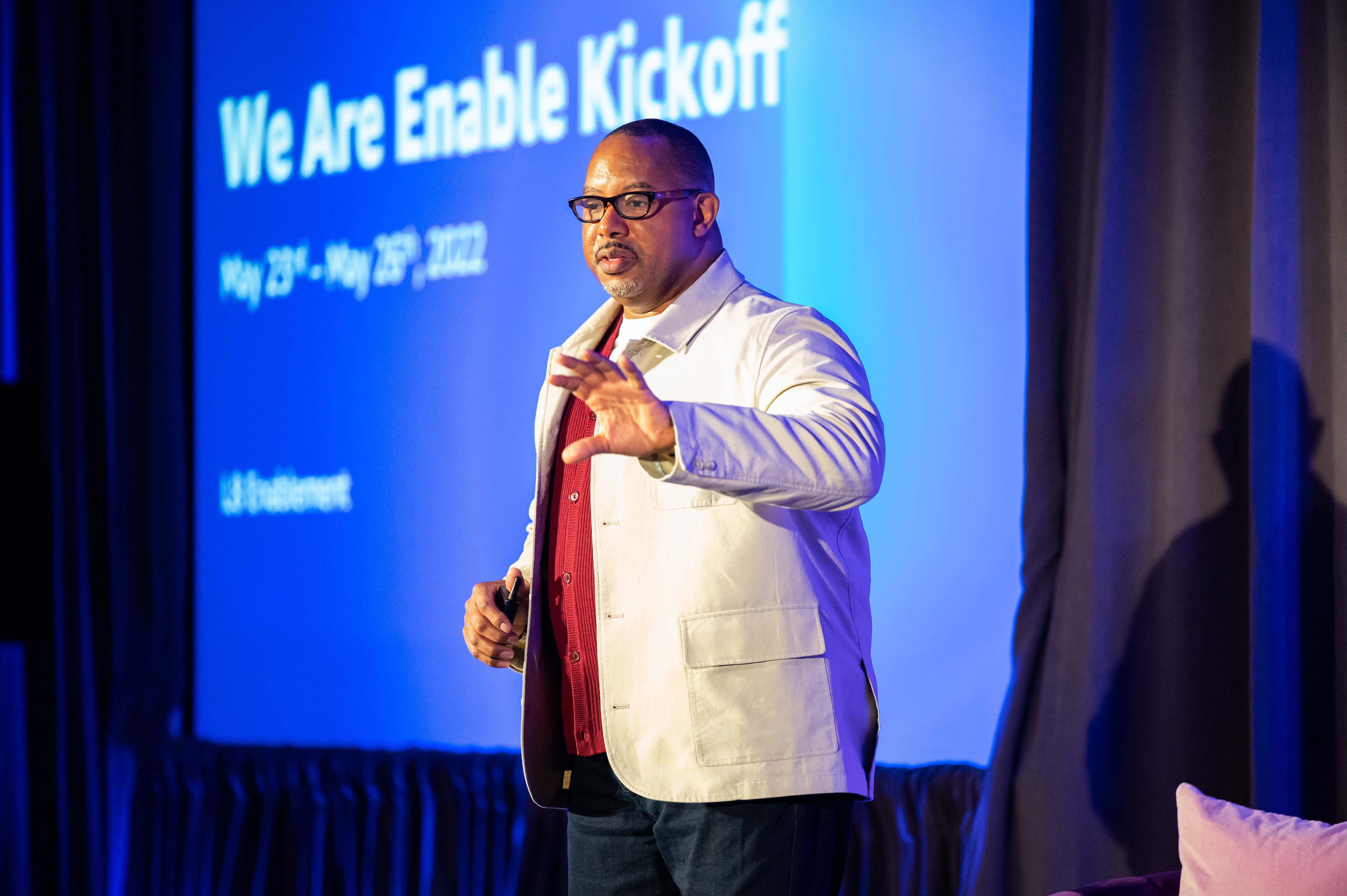 A speaker presents on stage in front of a blue screen that reads 'We Are Enable Kickoff, May 23rd - May 25th, 2022.'