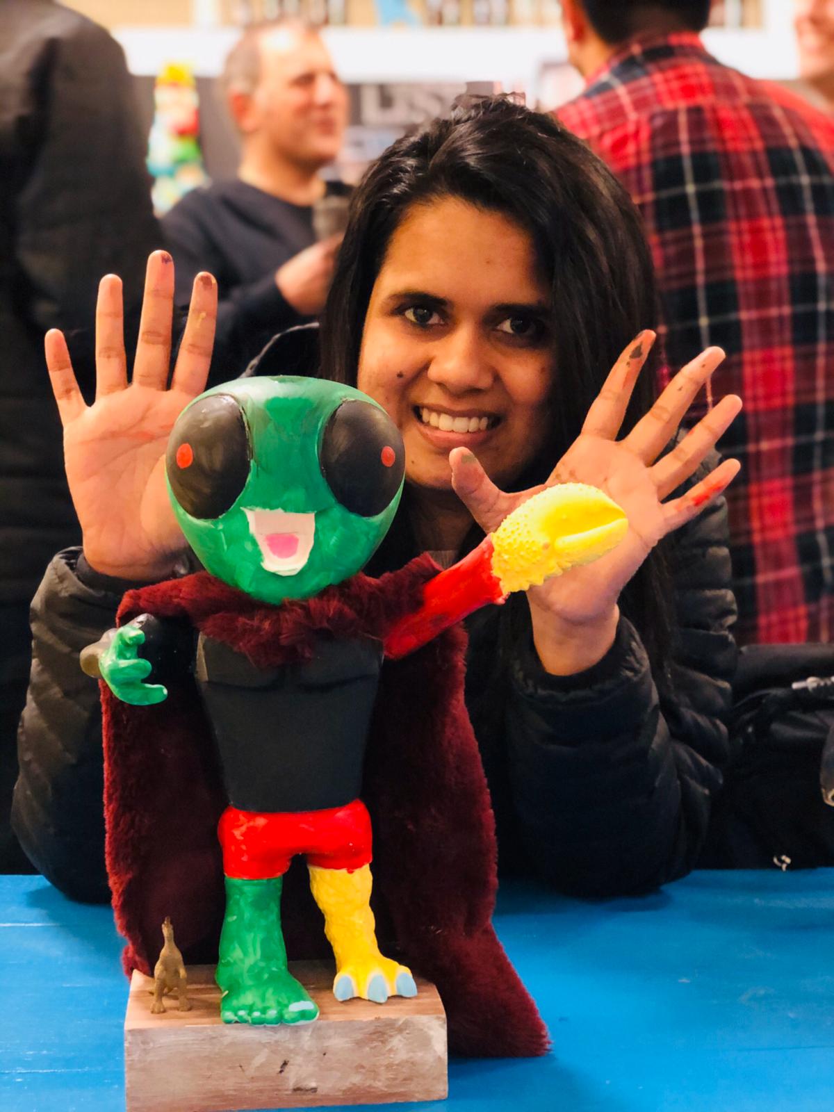 A woman with painted hands smiles behind a colorful green alien figure statue at an art event, with people in the background.
