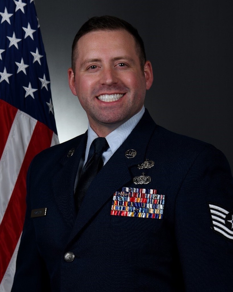 A smiling military service member in dress uniform stands in front of an American flag, with medals and insignias displayed on the uniform.