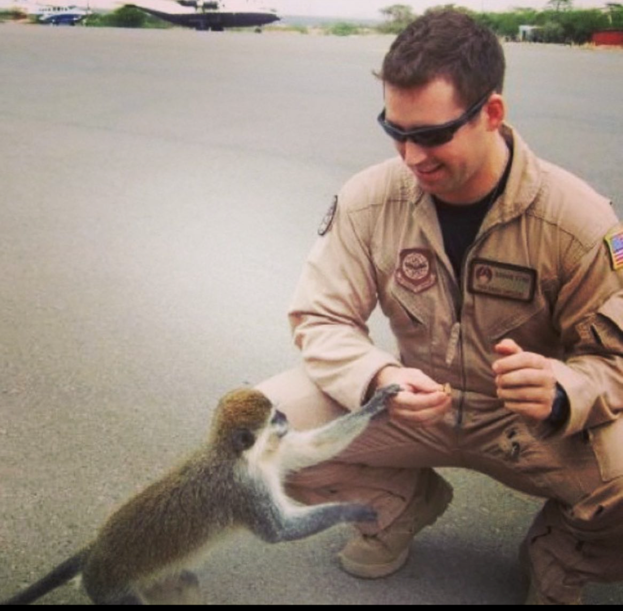 A person wearing a beige uniform and sunglasses kneels outdoors, interacting with a small monkey that is reaching out towards them.