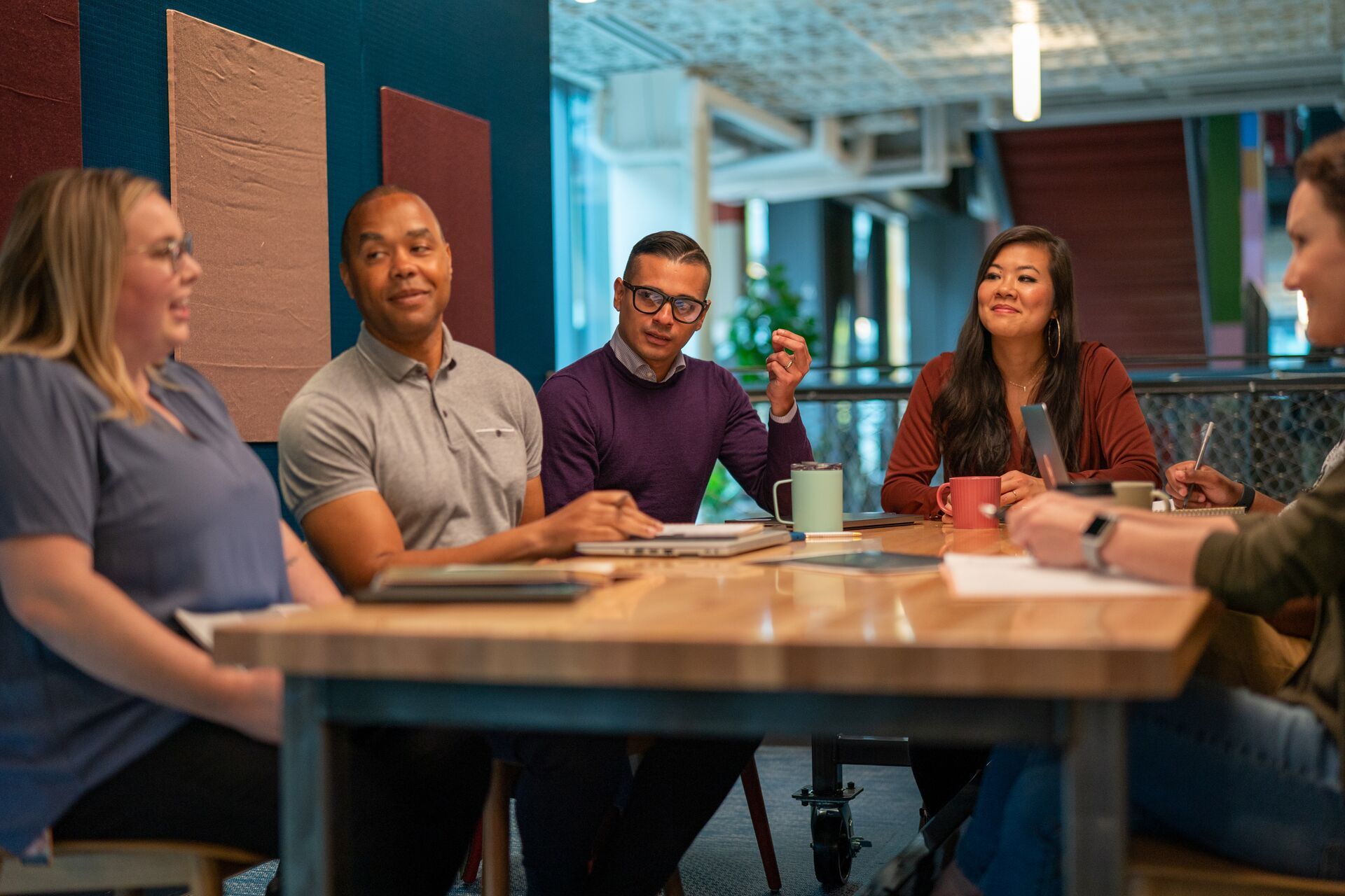 A group of diverse professionals are collaborating and discussing around a meeting table in a modern office environment. The team is engaged and working together, surrounded by notebooks, laptops, and coffee mugs.