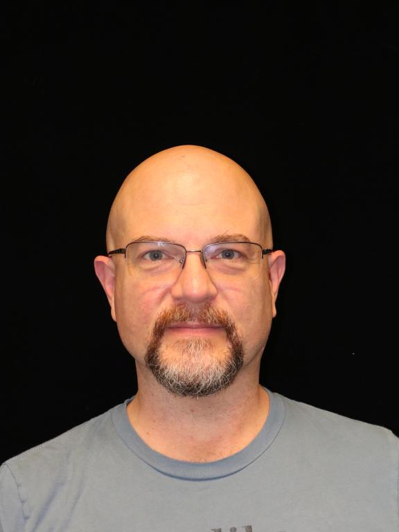 Portrait of a bald man wearing glasses and a goatee, facing forward against a black background.