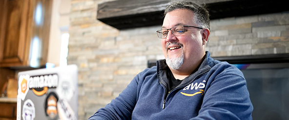 An AWS employee working remotely on a laptop, smiling at home with a laptop featuring various stickers, including an Amazon logo.