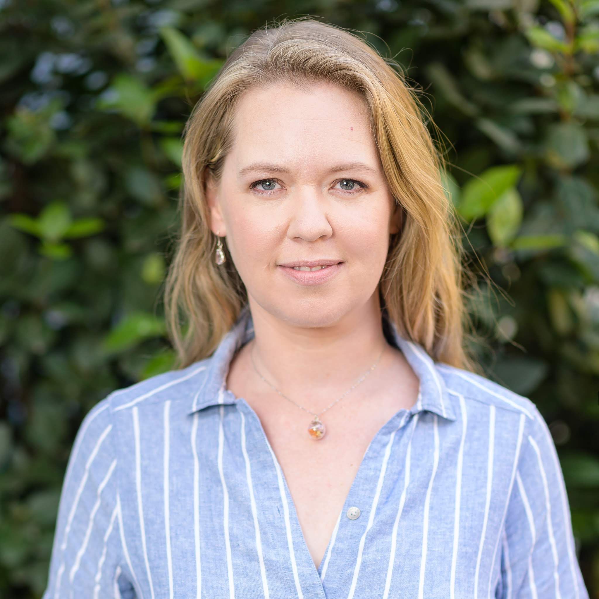 A professional outdoor portrait of a woman with blonde hair, wearing a blue and white striped shirt, standing in front of green leafy background.