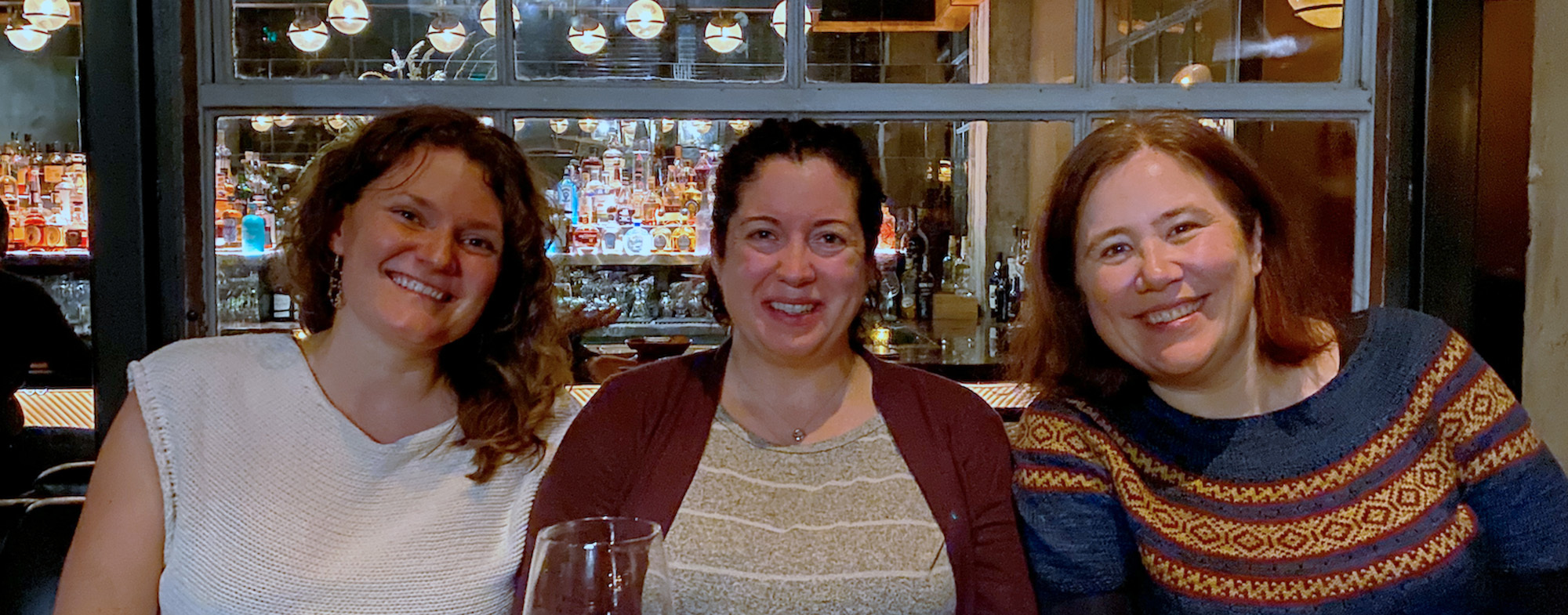 Three women smiling while sitting together at a dinner table with a bar background.