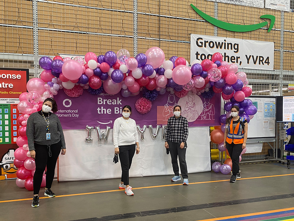 Four Amazon employees pose in front of a balloon arch and a 'Break the Bias' banner during an International Women's Day event at the YVR4 facility. The event backdrop is decorated with pink and purple balloons, and there are COVID-19 safety protocols in place with everyone wearing masks.