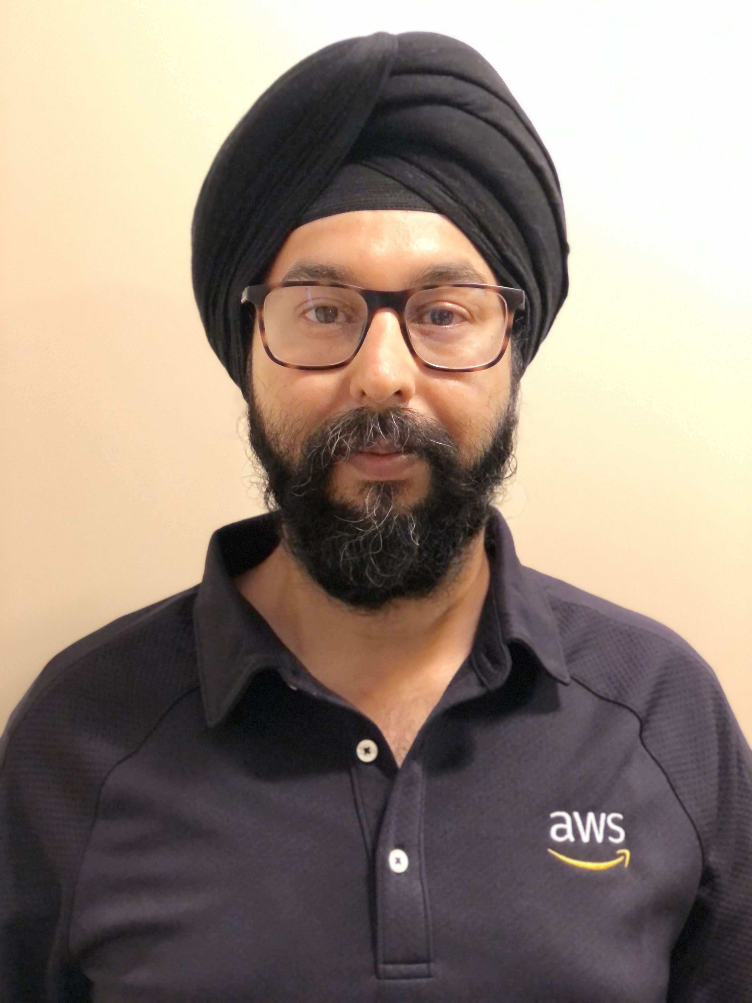 Portrait of an AWS employee wearing a black turban, glasses, and an AWS-branded shirt, photographed in front of a plain background.
