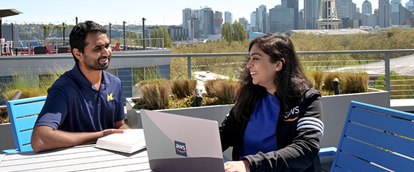 Two AWS employees have an outdoor meeting on a rooftop terrace with laptops and notebooks, overlooking the Seattle skyline in the background on a sunny day.