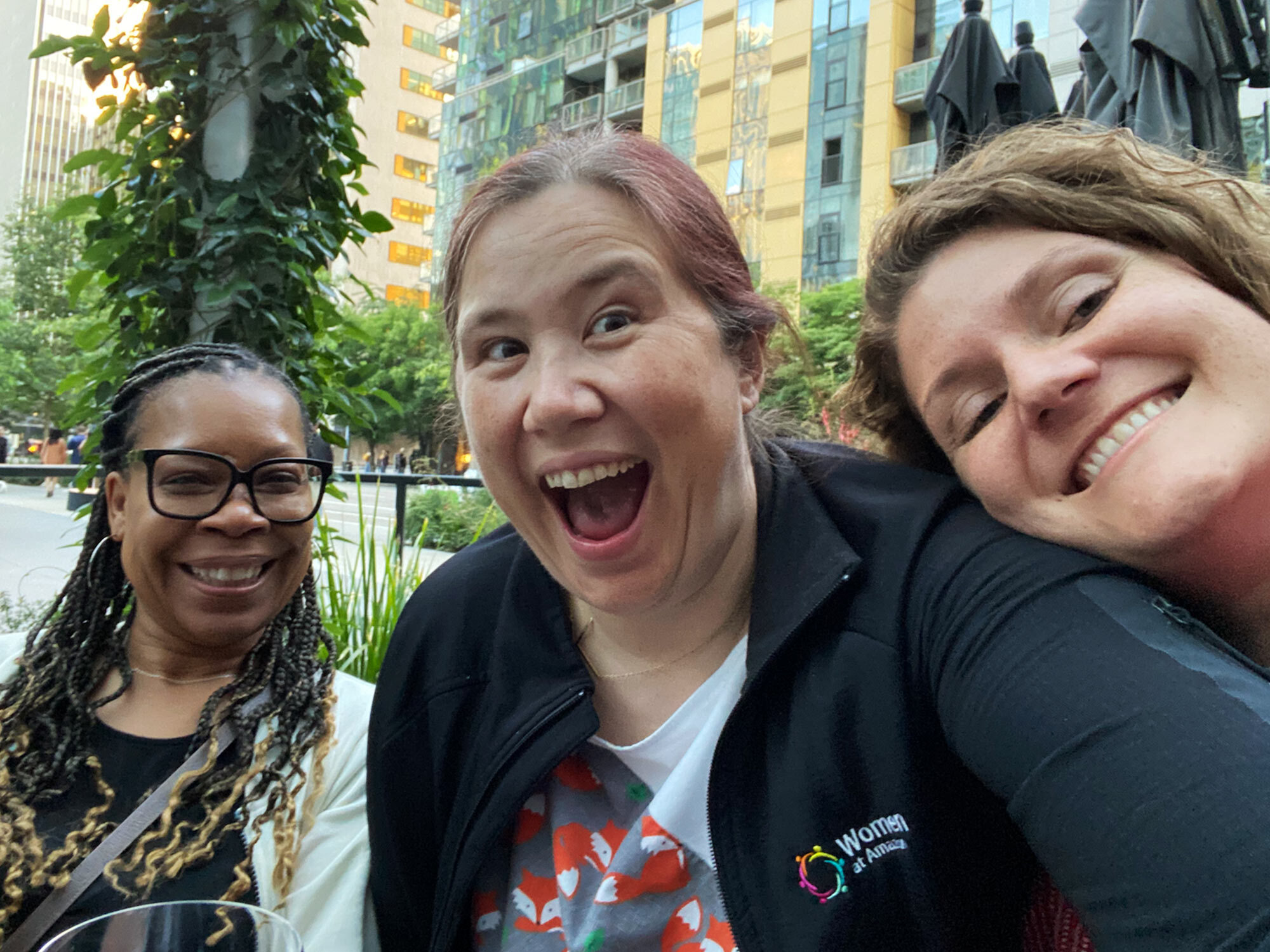 Three women smiling and taking a group selfie outdoors, with city buildings and greenery in the background. One person is wearing a 'Women at Amazon' branded jacket.