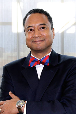 A professional business portrait of a man in a dark suit and a red and blue bowtie, standing confidently with his arms crossed.