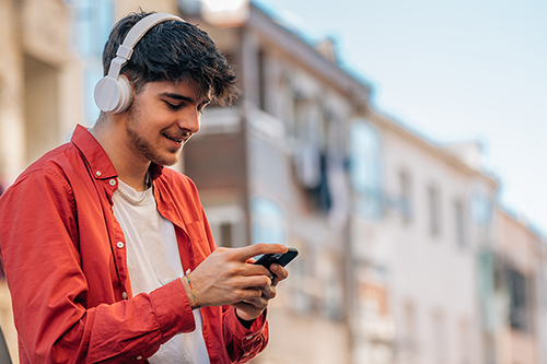 A young man wearing headphones is using a mobile phone while standing outdoors on a city street. He appears to be enjoying music or media, smiling and focused on his device.