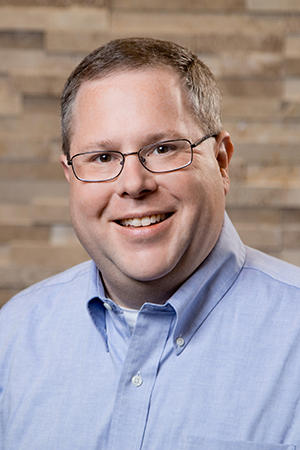 Professional headshot of a smiling man wearing glasses and a light blue shirt, posed in front of a brick wall background.