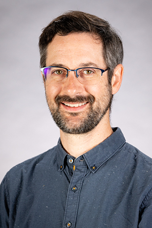 A man with short dark hair, glasses, and a beard is smiling at the camera. He is wearing a blue button-up shirt and is posed in front of a light gray background.