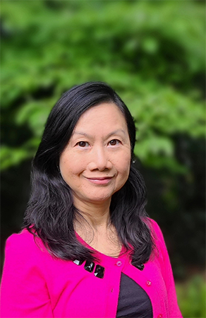 A woman with long dark hair, wearing a bright pink cardigan and dark top, poses outdoors in front of green foliage.