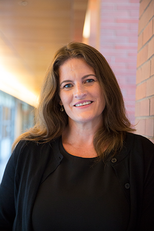 A professional indoor portrait of a woman with long hair, wearing a black top and standing against a brick wall, smiling at the camera.