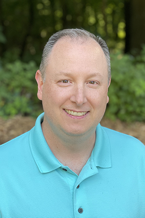 Outdoor portrait of a smiling person wearing a blue polo shirt, standing in front of greenery.
