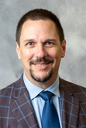 Professional headshot of a man in a checked suit jacket, blue shirt, and blue tie with a neutral background.