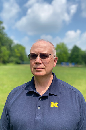 A person wearing sunglasses and a navy blue Michigan shirt stands outdoors in a grassy area under a partly cloudy sky.
