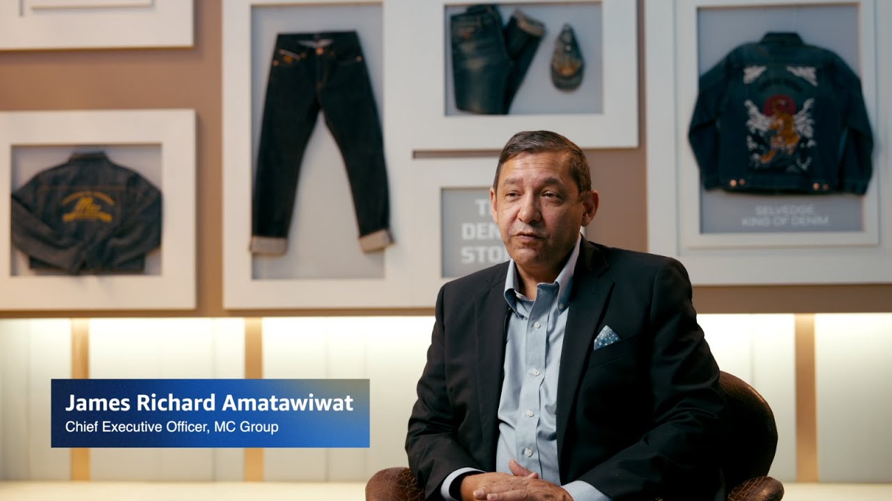 James Richard Amatawiwat, Chief Executive Officer of MC Group, is seated for an interview against a denim-themed background with framed jeans and jackets on the wall.