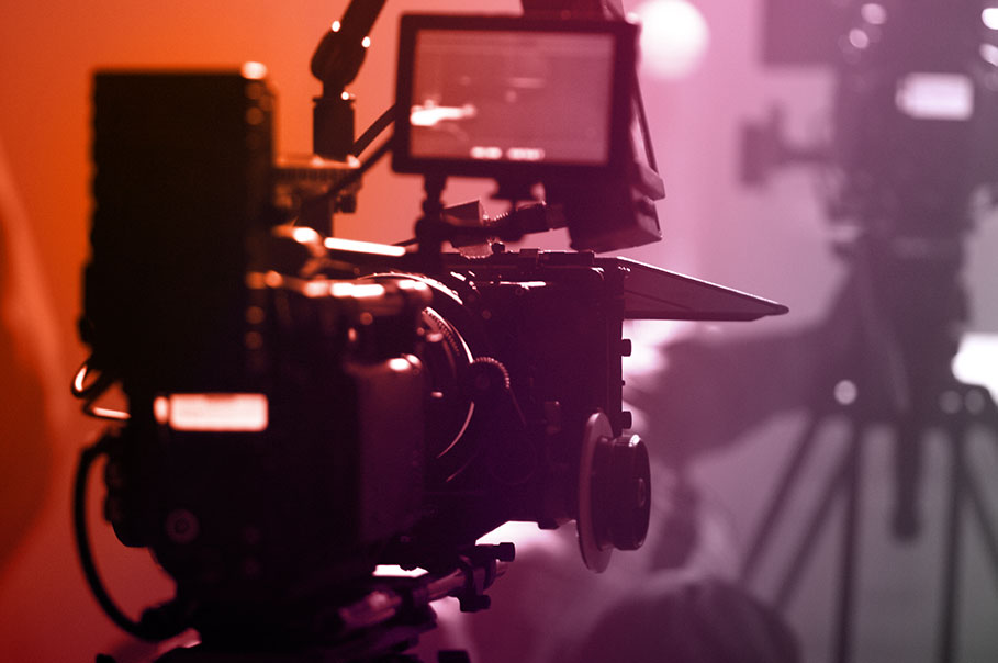 Close-up of a professional video camera on a set, with a warm orange and purple gradient lighting.