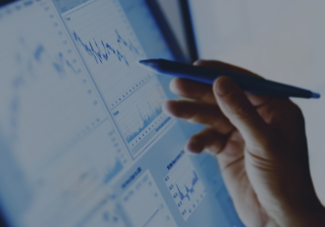A hand holding a pen points at a computer screen displaying financial graphs and charts.