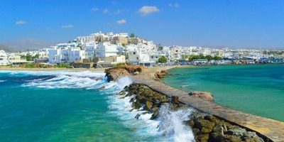 A scenic view of the Naxos coastline in Greece showing white buildings, a rocky pier, turquoise sea water, and the concept of a smart island.