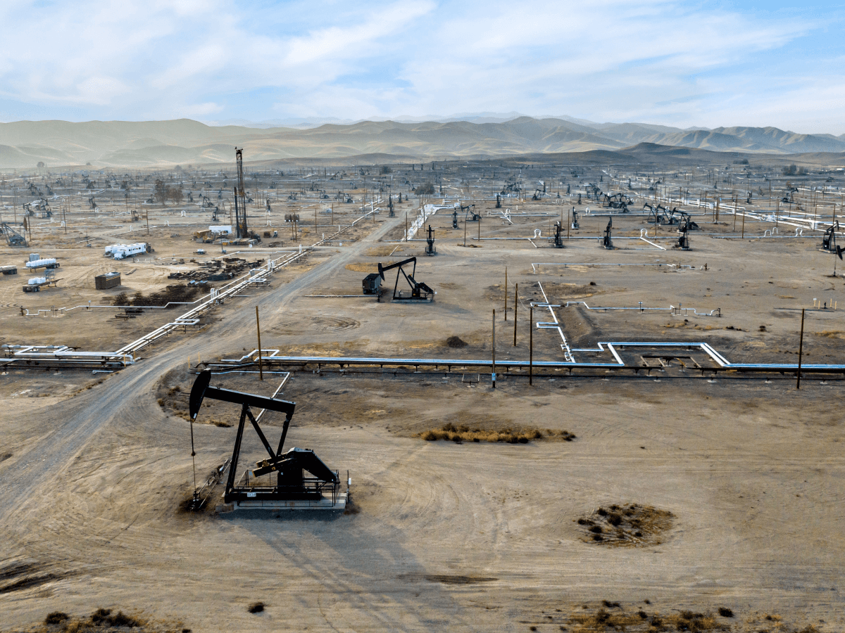 Aerial view of an expansive oil and gas field with numerous pumpjacks operating in a desert landscape with mountains in the distance.
