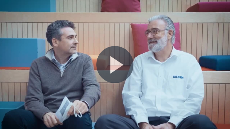 Two men sit on colorful bench seating, engaged in conversation. One wears a shirt with the 'Belden' logo, suggesting a discussion about modernizing global manufacturing operations, associated with Belden and TensorIoT.