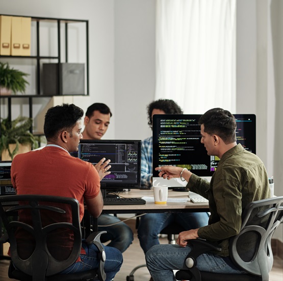 Group of people collaborating at a table with computer screens displaying code in a modern office setting.