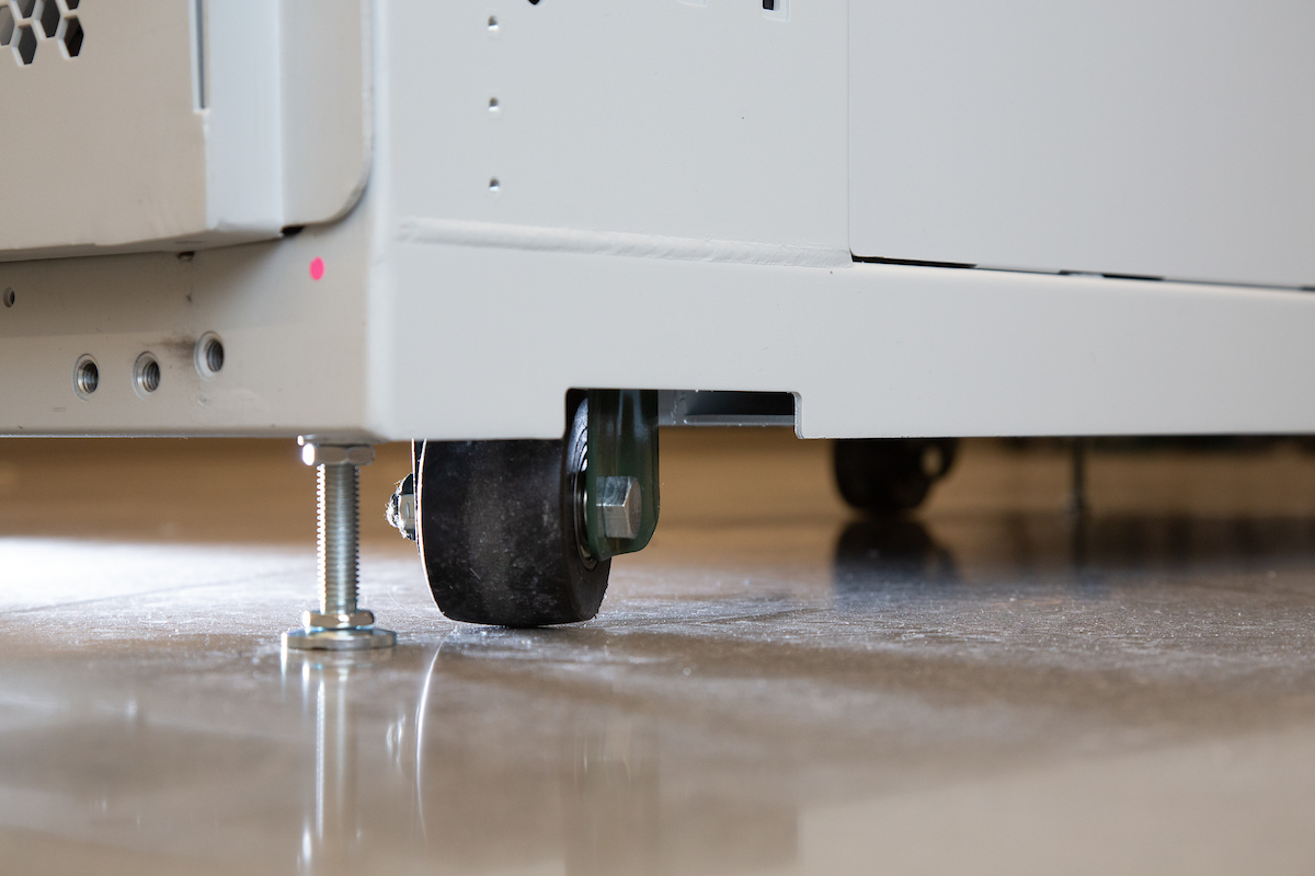 Close-up of a white metal cabinet base with a caster wheel and adjustable leveling foot on a reflective floor.